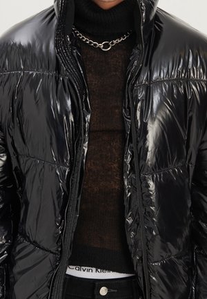 Mann in glänzender schwarzer Daunenjacke, schwarzem Rollkragenpullover, silberner Kette und sichtbarem Calvin Klein Bund.
