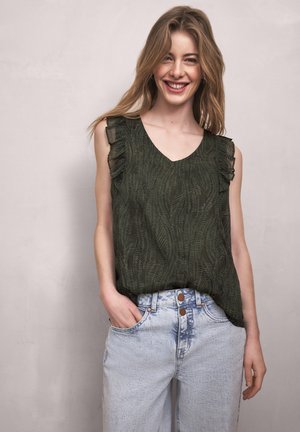 Uśmiechnięta kobieta ubrana w ciemnozieloną, bezrękawnikową bluzkę z falbanami oraz jasnoniebieskie, wysokiej talii dżinsy, stojąca na jednolitym tle.