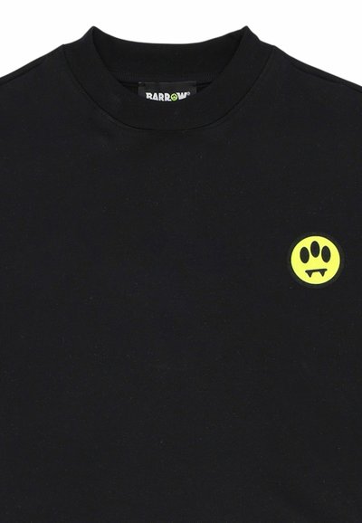 T-shirt noir à col rond avec un petit logo circulaire jaune sur la poitrine représentant une patte et des crocs de vampire.
