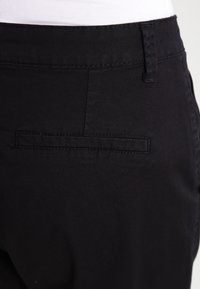 Pantaloni in tessuto nero con una tasca posteriore dotata di un pattina rettangolare e cuciture sottili. Il materiale appare liscio con una finitura opaca.