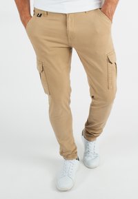 Pantalons cargo beiges avec poches latérales, en mélange de coton, coupe fuselée, associés à des baskets blanches. Design simple, sans motifs ni accents.