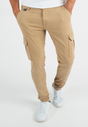 Pantalons cargo beiges avec poches latérales, en mélange de coton, coupe fuselée, associés à des baskets blanches. Design simple, sans motifs ni accents.