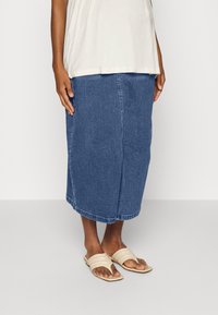 Saia midi de denim em azul profundo, com corte reto, costura frontal e bainha desfiada. Usada com sandálias beiges de tiras horizontais.