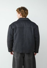 Jong persoon met kort, krullend haar draagt een donkergrijze gewatteerde jas met ruiten en losse zwarte spijkerbroeken, staande met de rug naar een witte muur.