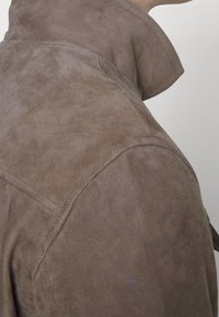 Ruskea mokkatakki, jossa on korkeakauluksellinen malli, tekstuuri pinta ja olkasaumadetailit. Ei näkyvää metalliosaa tai kuvioita.