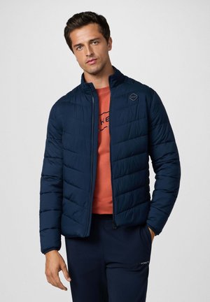 Marineblaue Steppjacke mit gestepptem Muster, hohem Kragen, Reißverschluss und elastischen Bündchen, über einem orangefarbenen Hemd getragen. Glatte Textur.