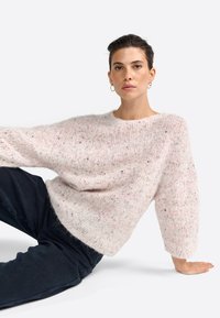 Helles rosa Strickpullover mit lockerer Passform und strukturiertem Stoff, der Farbspritzer aufweist. Lange Ärmel und weiter Ausschnitt. Getragen mit dunklen Hosen.