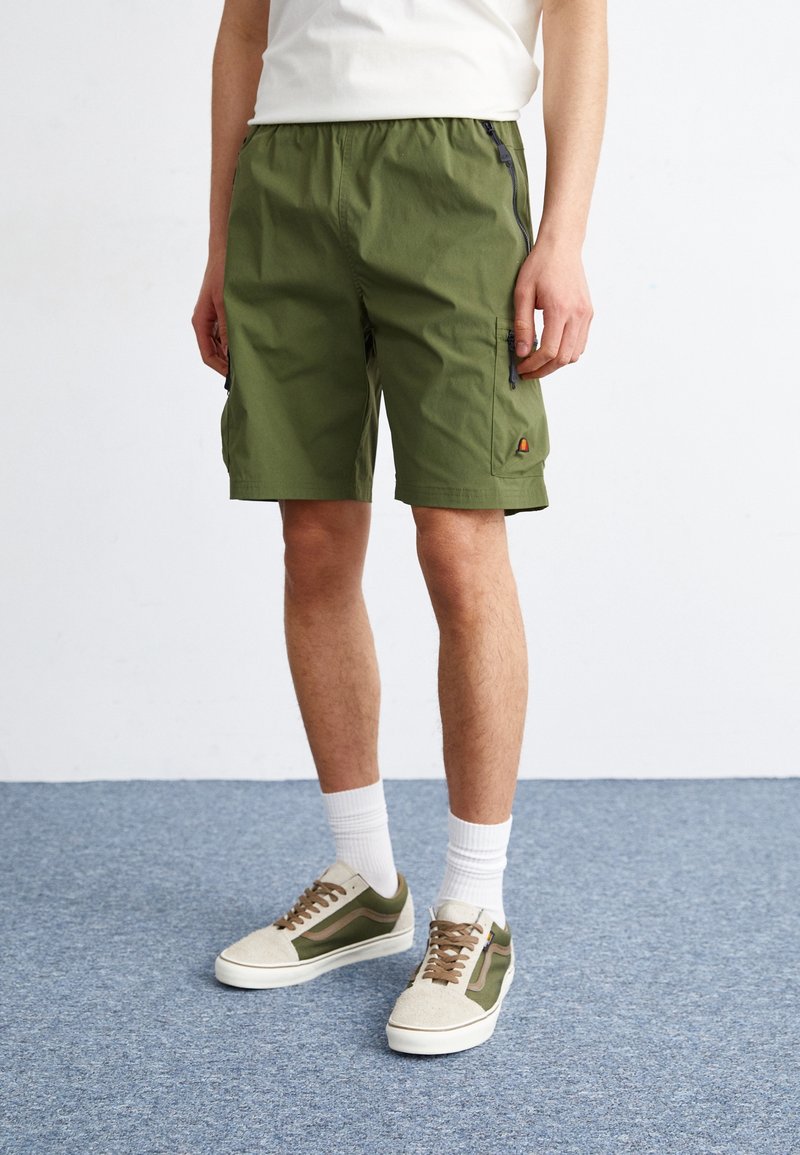 Shorts vert olive avec une taille élastique, poches latérales avec zippers et un petit logo en accent. Associés à des baskets beiges et vertes.