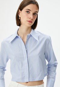 Camicia abbottonata cropped di colore azzurro chiaro con righe bianche verticali. Presenta un colletto a punta e maniche lunghe con una texture di tessuto liscia.