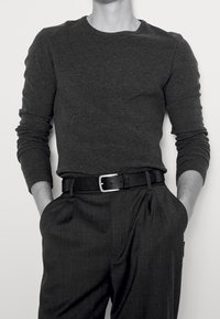 Une chemise grise à manches longues avec un col rond, associée à un pantalon plissé anthracite et une ceinture tressée noire avec une boucle rectangulaire en argent.