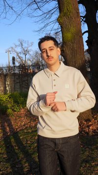 Νέος άνδρας με μπεζ πουλόβερ με μακριά μανίκια και σκούρο τζιν, που στέκεται έξω δίπλα σε ένα μεγάλο δέντρο με γυμνά κλαδιά σε μια ηλιόλουστη μέρα.