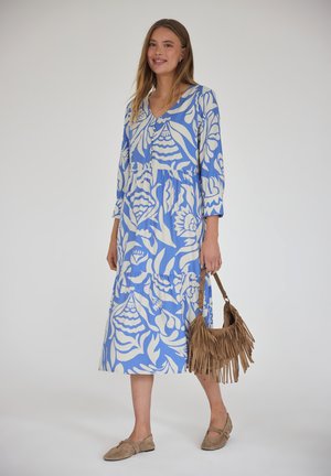 Kobieta w niebiesko-białej wzorzystej sukience midi, trzymająca brązową torebkę z frędzlami i nosząca beżowe płaskie buty, stojąca na białym tle.
