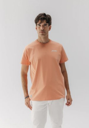 Camiseta de algodón color durazno con cuello redondo, que presenta un pequeño logo blanco en el pecho y un acento azul en el dobladillo.
