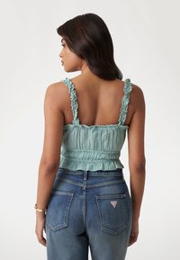 Light green crop top with ruffled straps, smocked texture, and a flared hem, paired with high-waisted denim jeans featuring a logo patch.