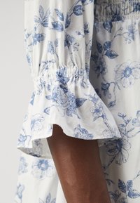 Vit klänning i tyg med blå blommönster. Har samlade ärmar och utsvängda manschetter som visar detaljerade blom- och lövmönster.