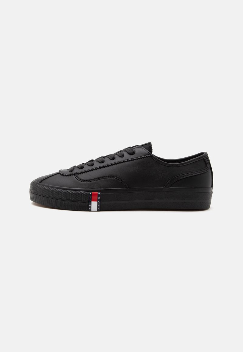 Μαύρο sneaker χαμηλού τύπου με μαύρα κορδόνια και ένα μικρό ορθογώνιο κόκκινο, λευκό και μπλε λογότυπο που γράφει "Tommy Jeans" στην πλευρά της σόλας.