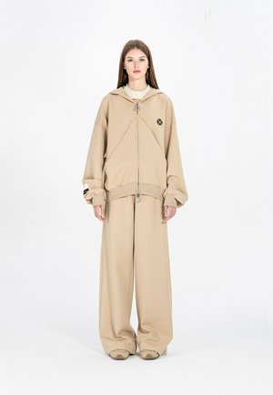 Joven mujer está de pie mirando hacia adelante, con una sudadera beige de gran tamaño con cremallera frontal y pantalones anchos a juego y zapatillas deportivas.