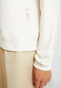 Primer plano de una mano al lado de pantalones beige y una camisa de manga larga color blanco roto con el texto rojo "LOVE" y un corazón bordado cerca de la costura lateral.