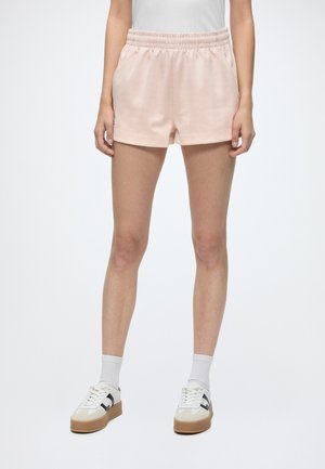 Personne portant un short taille élastique rose clair, des chaussettes blanches et des baskets blanches avec des accents noirs sur un fond uni.
