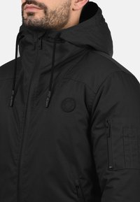 Black insulated jacket with a hood, featuring a zip front, two side pockets, and a patch logo on the chest. Smooth texture, sturdy material.