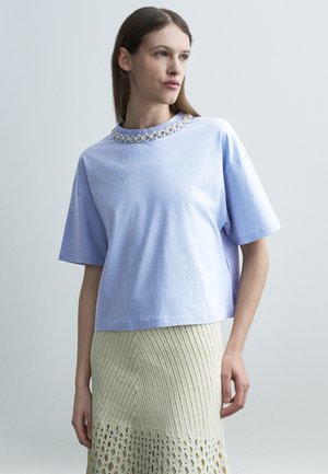 Kvinne iført en lys lilla kortermet topp med perlepynt på kragen og et kremfarget strikket skjørt med et teksturert mønster.