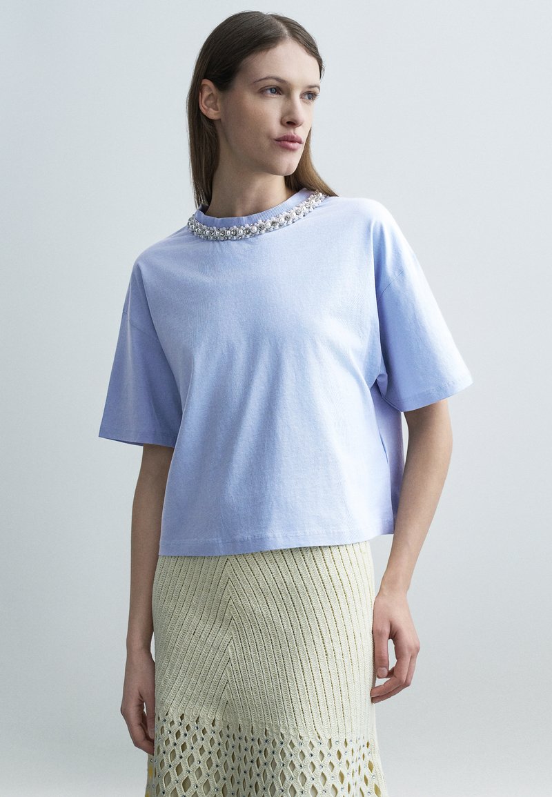 Γυναίκα που φοράει ανοιχτό μωβ κοντομάνικο μπλουζάκι με διακοσμητικά μαργαριταρένια στοιχεία στον γιακά και κρεμ πλεκτή φούστα με υφή.