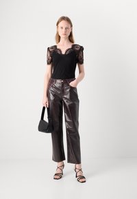 Haut noir avec des manches en dentelle, associé à un pantalon marron brillant taille haute, un sac à main noir et des sandales noires à lanières. Fond simple.