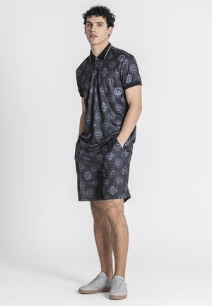 Mand står med hænderne i lommerne iført matchende sort mønstret polo shirt og shorts med lysegrå sneakers.