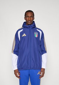Modrá vetrovka s bielymi rukávmi, zlatými pruhmi adidas a emblémom Talianska. Má kapucňu a predné zapínanie na zips.