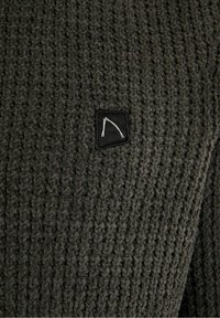 Gestrickter Pullover in Dunkelgrau mit strukturiertem Muster, versehen mit einem quadratischen schwarzen Patch mit weißem Logo. Das Material wirkt weich und dick.