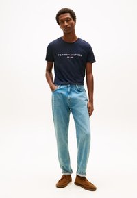 Mies, joka on pukeutunut laivastonsiniseen Tommy Hilfiger -t-paitaan, vaaleansinisiin farkkuihin ja ruskeisiin kenkiin, seisoo yksinkertaista valkoista taustaa vasten.