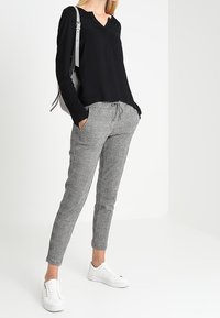 Un haut noir à manches longues avec un décolleté en V, associé à un pantalon gris à carreaux et des baskets blanches ; un sac à main gris complète la tenue.