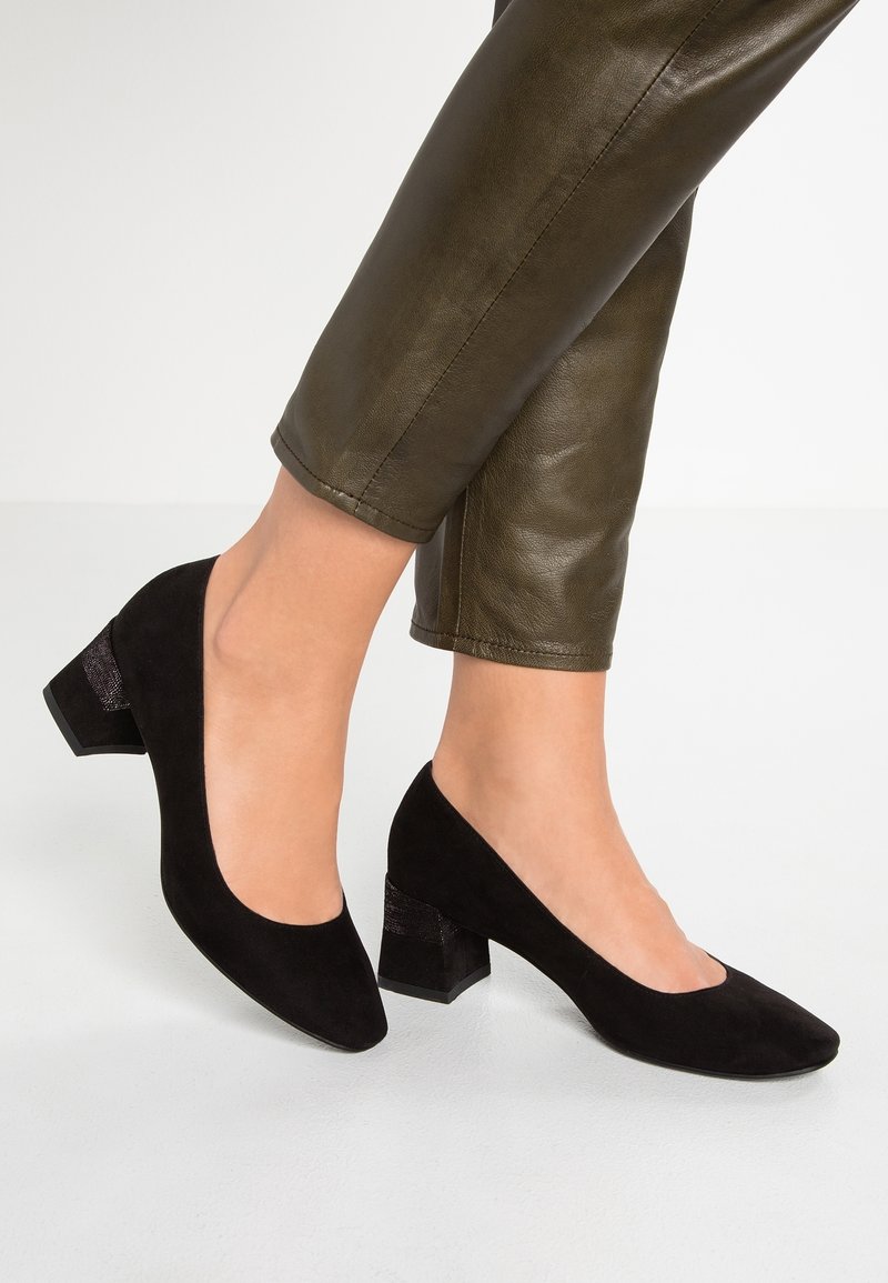 Jambes portant un pantalon en cuir vert olive et des chaussures à talons carrés en daim noir, debout sur un sol blanc.
