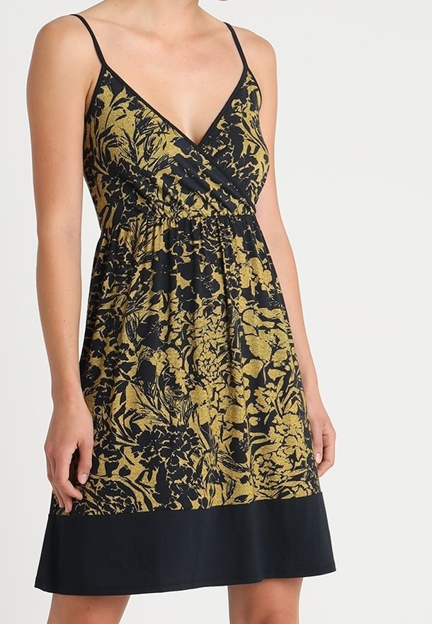 Femme portant une robe sans manches noire avec un motif floral doré, décolleté en V et une bande noire ourlée en bas.