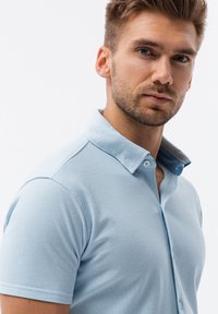 Camisa polo azul claro con un acabado texturizado, de mangas cortas y con un cuello a contraste. Presenta cierre de botones y un diseño ajustado.