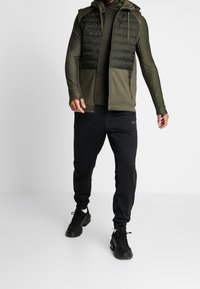 Uomo che indossa un gilet trapuntato verde oliva sopra una camicia a maniche lunghe con texture, pantaloni jogging neri e sneakers nere, in piedi su uno sfondo bianco.
