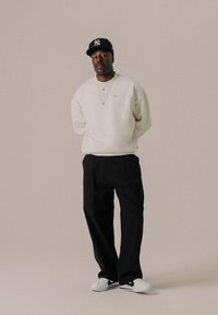 Crème sweatshirt met gouden logo, losse zwarte broek en witte sneakers met zwarte accenten. Accessoires omvatten gelaagde kettingen en een zwarte pet.