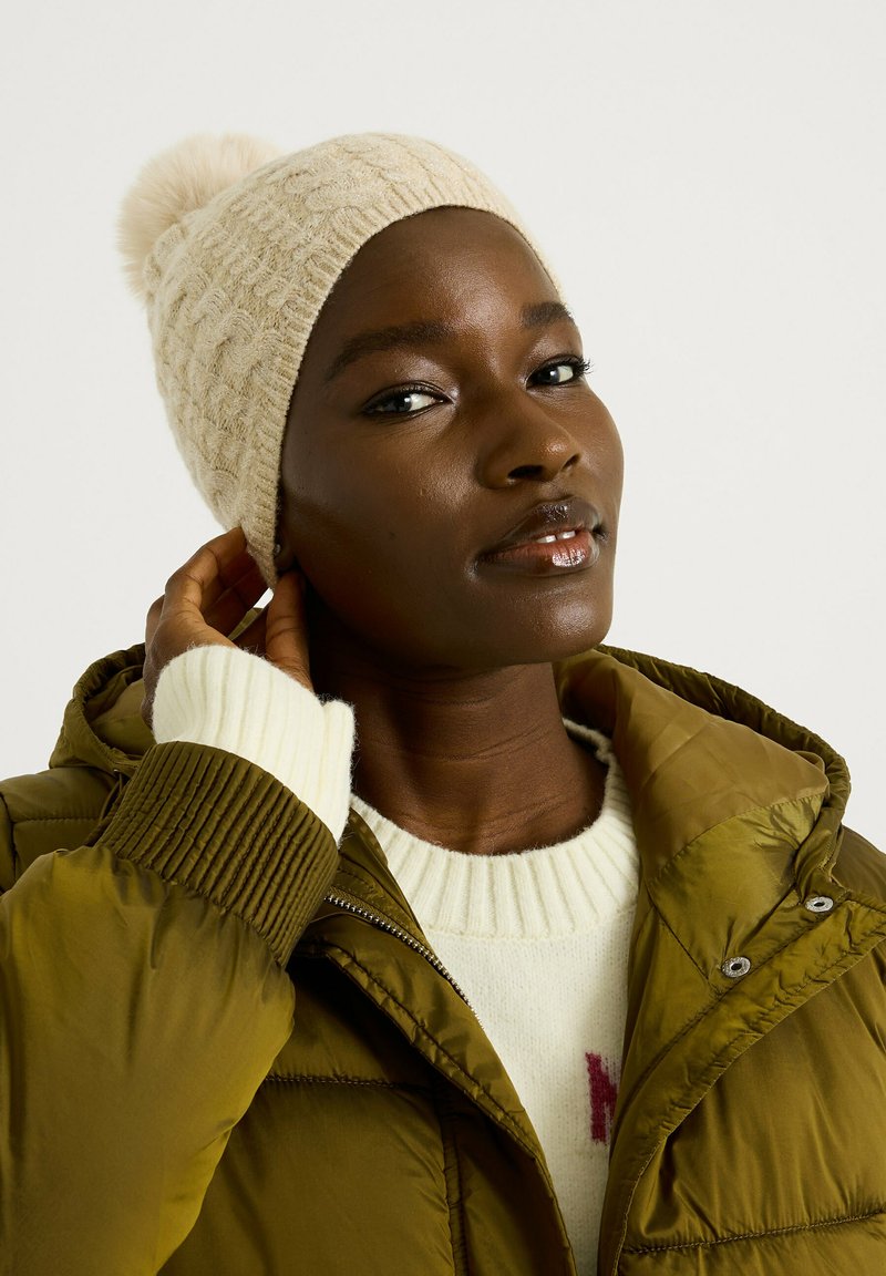 Bonnet en tricot crème avec un pompon sur le dessus. Le modèle porte une veste matelassée verte olive et un pull blanc avec des poignets côtelés et un motif rouge.