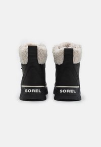 Sorte ankelstøvler i svart med en plysj, kremfarget pelskrage, gummisåle og hvit "SOREL"-merkevare på hælen. Teksturert skinnmateriale.