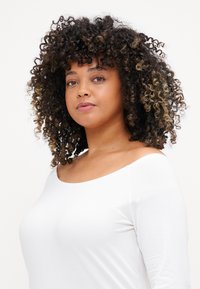 Top blanco de hombros descubiertos con mangas largas. El modelo tiene el cabello rizado, de longitud hasta los hombros, con mechas más claras.