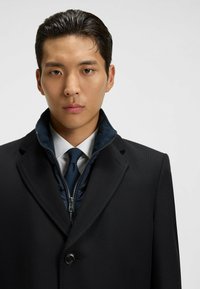 Cappotto in misto lana nero con colletto a scollo a V, chiusura a doppio petto con bottoni e una giacca interna blu navy con zip visibile.