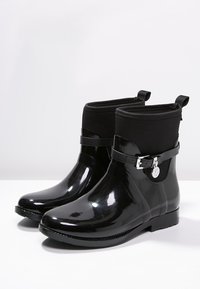 Une paire de bottes de pluie noires brillantes à cheville avec des poignets en tissu, des sangles à boucle sur le côté et des charms en forme de logo rond sur une surface blanche.