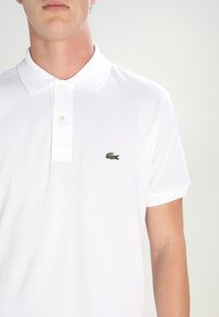 Chemise polo blanche en coton texturé, avec un col classique et des manches courtes. Logo crocodile vert brodé notable sur la poitrine gauche.