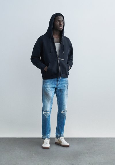 Νεαρός άνδρας που φοράει μαύρο φούτερ με φερμουάρ, λευκό αμάνικο μπλουζάκι, φθαρμένο τζιν μπλε και λευκά αθλητικά, στέκεται με τα χέρια στις τσέπες.