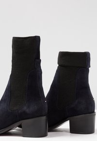 Paire de bottines en suède noir avec panneaux latéraux élastiques et talons blocs sur fond blanc.