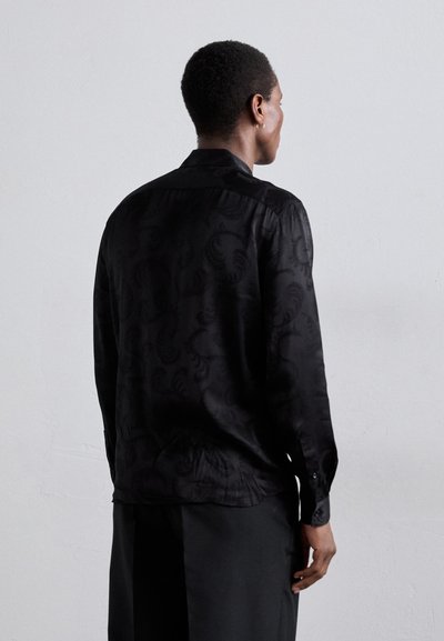 Camicia in satin nero con un sottile motivo a rilievo, maniche lunghe, colletto e polsini abbottonati. Indossata con pantaloni larghi.