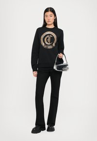 Schwarzer Pullover mit goldener kreisförmiger Logo, kombiniert mit schwarzen Schlaghosen und schwarzen klobigen Stiefeln; hält eine kleine silberne Handtasche.