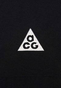 Črna tkanina z diagonalo bež vezenim logotipom, ki vsebuje črki "c" in "g", kar ustvarja močan kontrast in jasen dizajn.