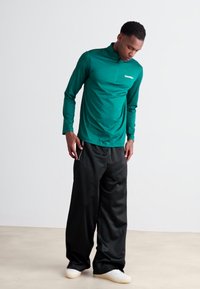Maglia a maniche lunghe, di colore teal, in tessuto tecnico con colletto con zip e logo, abbinata a pantaloni neri larghi e scarpe da ginnastica di colore chiaro.