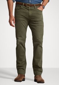Chinos de color verde oliva, tejido de algodón, diseño de pierna recta, cinco bolsillos, cinturón de cuero marrón con hebilla de metal, llevados con botas de ante marrón.